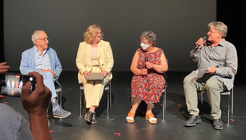 The speakers are each sitting on a chair in the middle of the space and facing the audience. Darrin Hagen is holding the microphone and talking to the three speakers.