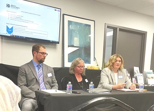 The speakers are sitting in front of the room and facing the audience. Three speakers are at one table with Kathryn Oviatt, K.C. discussing her presentation, which appears on the screen behind them.